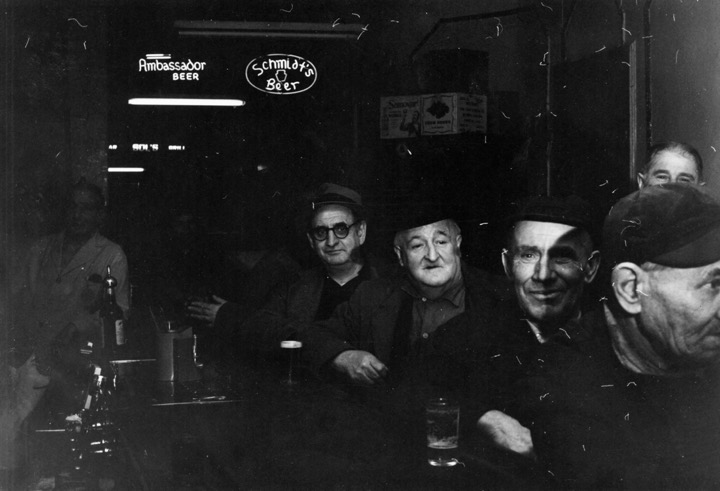 Black & white photo of several white men sitting at a bar, under neon signs for Ambassador and Schmidt's beers