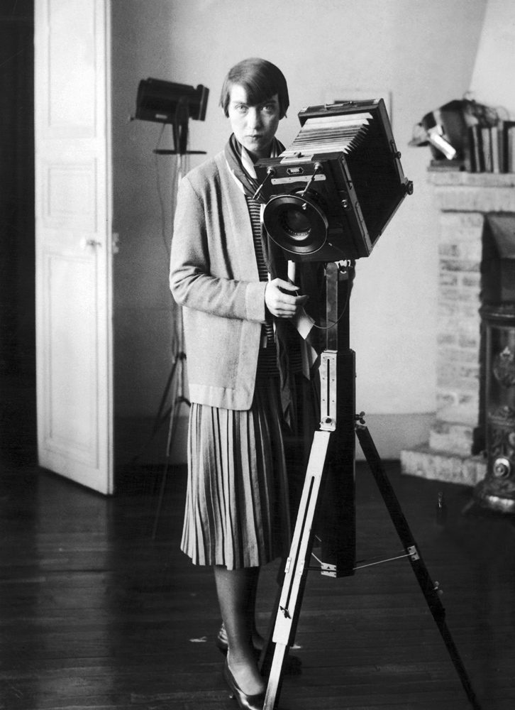 Abbott standing in a studio with a large camera, hair bob and gazing seriously at the viewer.