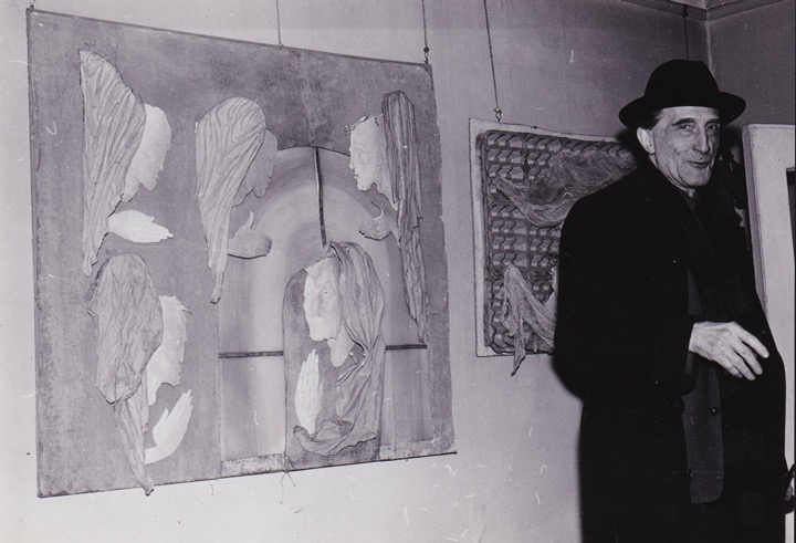 Duchamp smiles, as if mid speech, next to cardboard composition displaying the heads, hands, and wings of five angels surrounding an arched window