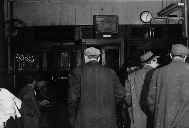 Several white men in hats and coats shown from behind leaving bar, TV and clock visible on wall above door.